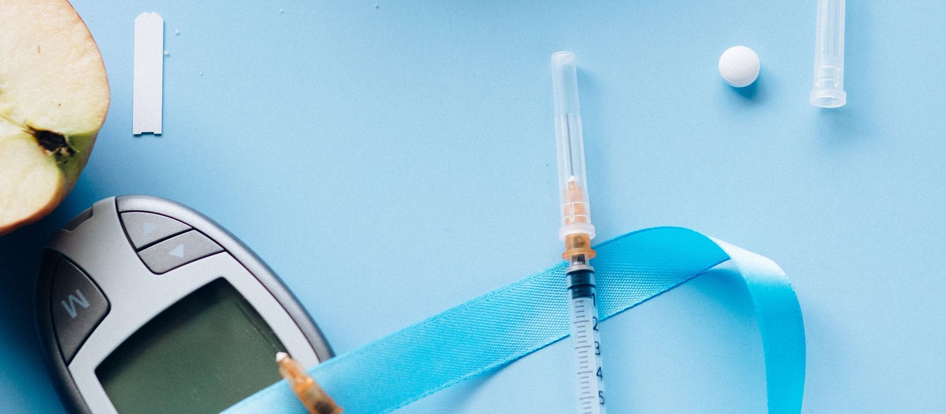 A glucose meter, a lancet and a sliced apple laid out on blue with the diabetes ribbon